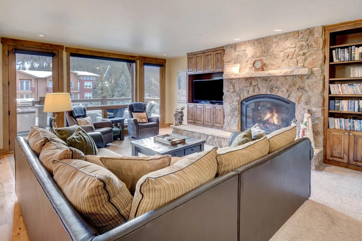 A cozy, expansive living room featuring a large sectional sofa centered around a stone fireplace, with built-in bookshelves and panoramic windows that flood the room with natural light and offer stunning mountain views. The space is styled with inviting, designer furnishings and is perfect for gatherings, relaxation, and movie nights.