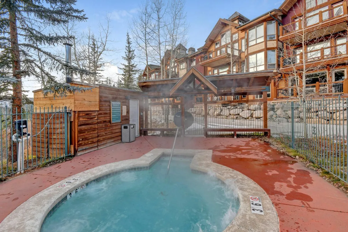 A tranquil exterior setting showcasing a community hot tub nestled among wooden structures and surrounded by mature trees, with the building visible nearby. The steam rising from the hot tub and the inviting seating area evoke relaxation and après-adventure comfort.