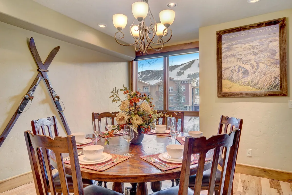 A cozy breakfast nook anchored by a dining table with a floral centerpiece, illuminated by a chandelier. Large windows offer natural light and picturesque views, enhancing the space's warmth and inviting atmosphere.