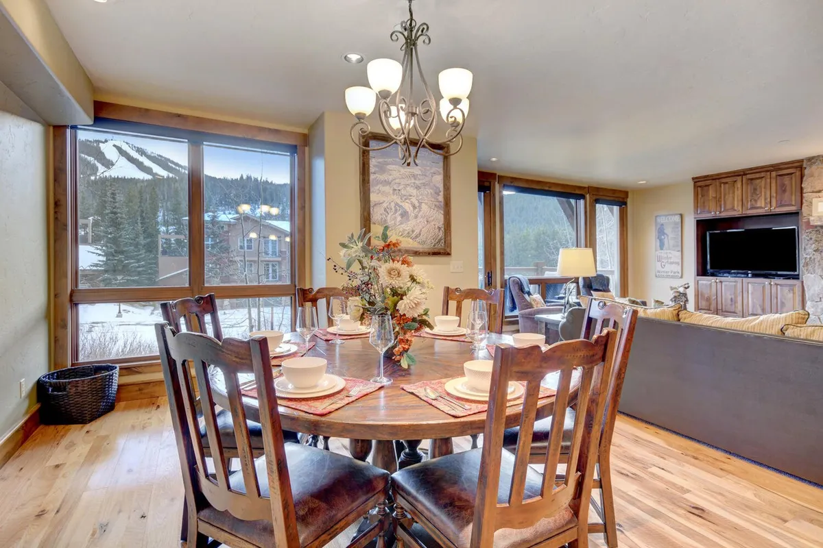 A stylish dining area with a large table that comfortably seats nine, illuminated by a modern chandelier. The space is defined by large windows framing panoramic mountain vistas, creating a warm, celebratory atmosphere suitable for festive family meals or casual breakfasts.