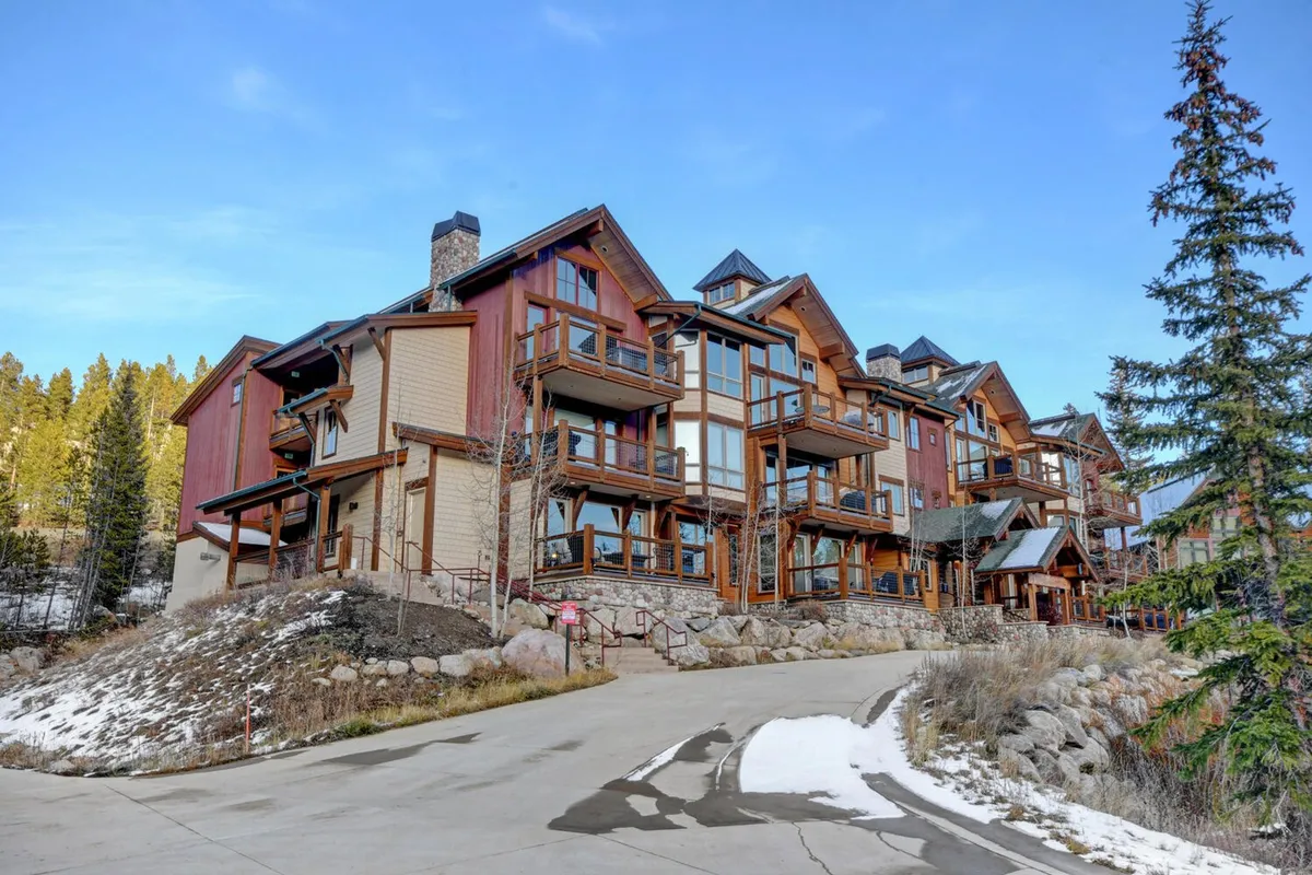 The hero image showcases a beautiful single-family mountain villa with rustic wooden balconies, elegant stone accents, and expansive windows. The home is set against a breathtaking mountain backdrop, exuding a warm, cozy atmosphere. Visible balconies invite guests to relax and take in the views, while the outdoor setting hints at activities like sunset watching and gathering around a fire pit, connecting directly to nature's beauty.