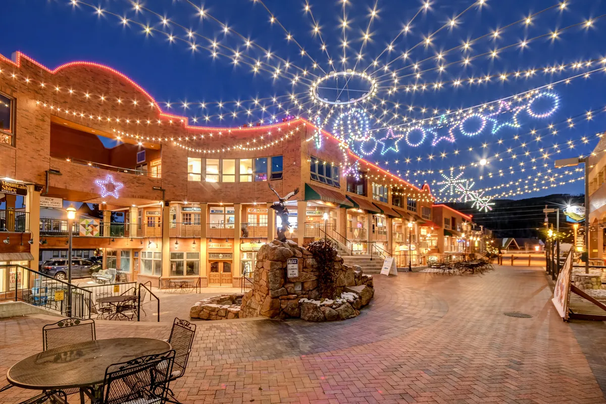 A welcoming village scene featuring storefronts, decorative string lights, a brick walkway, and a central fountain sculpture. The ambiance is lively and festive, suggesting an active downtown with dining, shopping, and social gatherings.