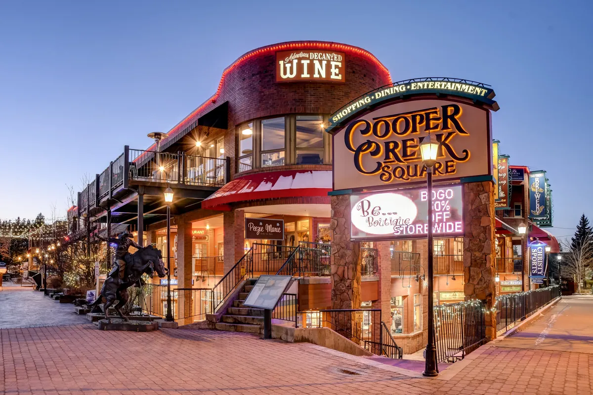 A lively exterior scene showcasing the Cooper Creek Square sign, an inviting wine shop, outdoor seating, and decorative lighting that fosters a festive, vibrant community atmosphere. The setup suggests opportunities for dining, shopping, and engaging in local events.