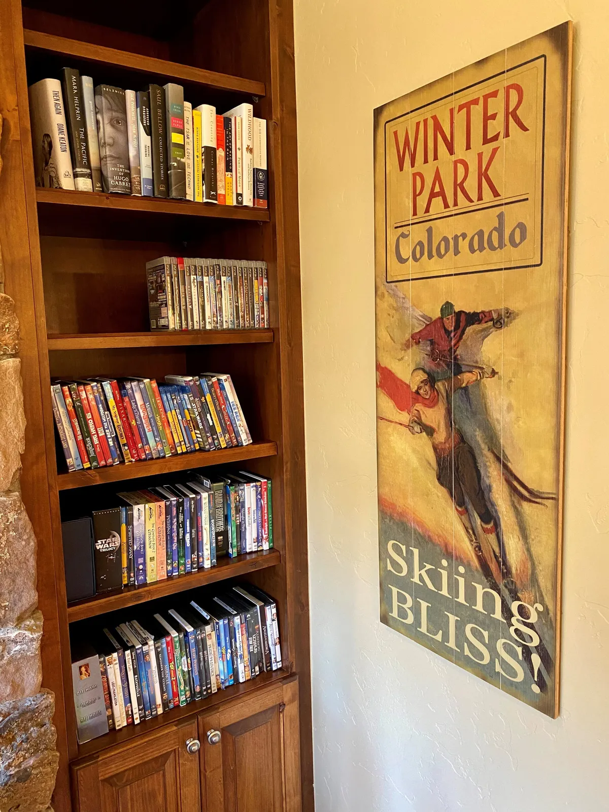 A warmly lit space featuring a wooden bookshelf stocked with books and DVDs, alongside a cozy seating area. Vintage ski decor and a nostalgic mood invite guests to enjoy board games or movie nights, adding to the entertainment options highlighted in the section.