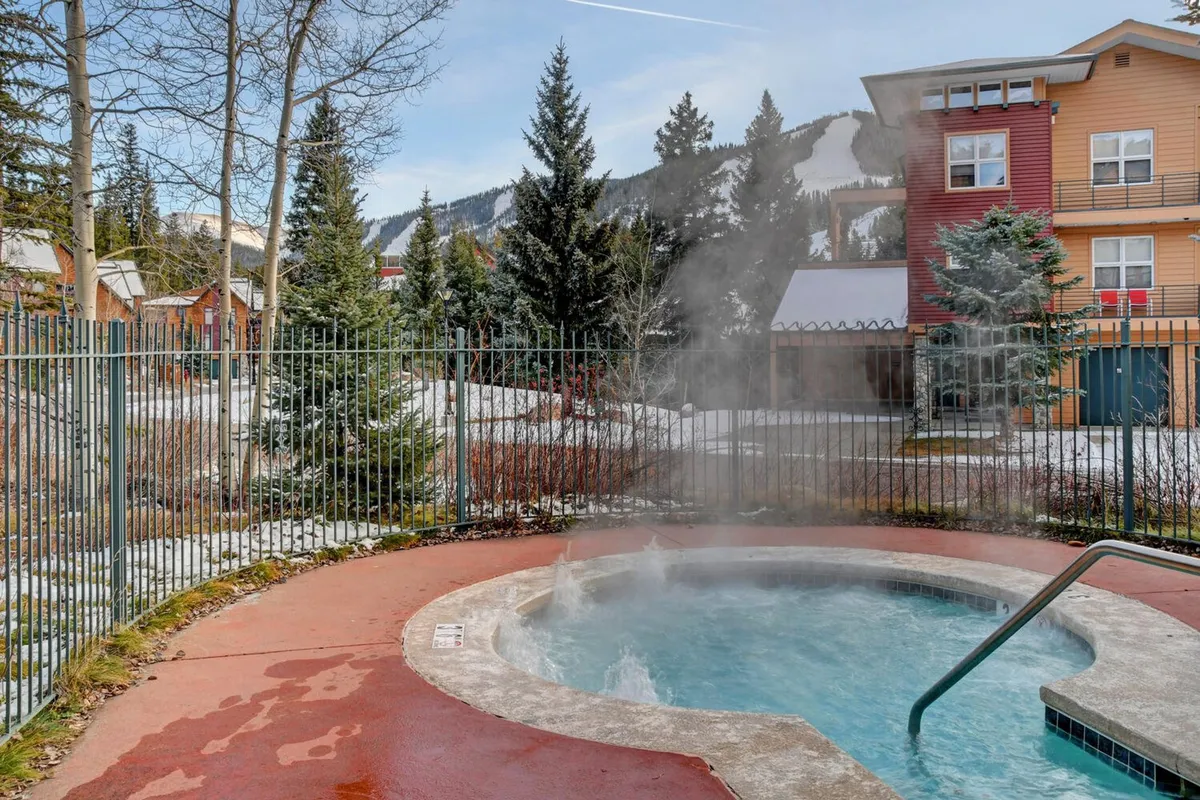 A serene outdoor community hot tub nestled among trees, with a panoramic mountain view in the background. Guests are shown relaxing and enjoying the tranquil atmosphere, ideal for après-ski soaks and socializing.