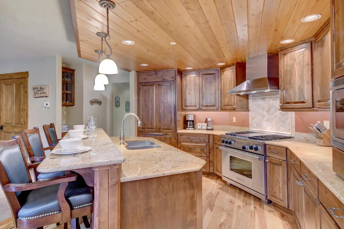 A welcoming kitchen scene featuring a prominent kitchen island with bar stools, warm wooden cabinetry, stainless steel appliances, and slab granite countertops. The space is designed for both cooking and entertaining, with ample workspace and an inviting atmosphere.